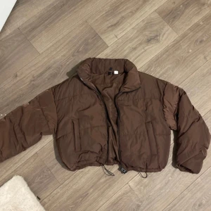 Brun pufferjacka - Säljer en brun pufferjacka med dragkedja och stora fickor. Jackan är tillverkad i 100% polyester och har en croppad passform. Perfekt för kyligare dagar. Alldrig använd