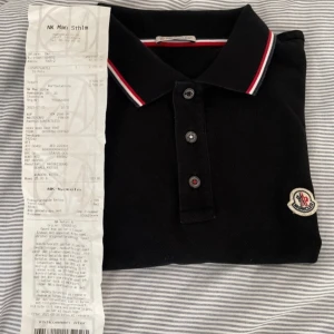 Svart pikétröja från Moncler - Säljer en snygg svart pikétröja från Moncler. Den har en klassisk krage med röd och vit randig detalj och tre knappar framtill. Tröjan är i nyskick och perfekt för både vardag och fest. Passar bra till jeans eller chinos för en stilren look.