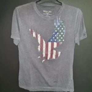 Grå t-shirt med amerikansk flagga och örn från Denim Day - Säljer en grå t-shirt från Denim Day i storlek medium. Den har en cool design med en örn som är fylld med den amerikanska flaggan på framsidan. T-shirten är i bomull och har korta ärmar. Den är lite sliten med några små fläckar och ett litet hål på ryggen, men fortfarande snygg och bekväm att ha på sig. Perfekt för en avslappnad stil!