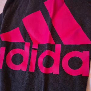 Svarta mjukisbyxor från Adidas - Säljer ett par svarta mjukisbyxor från Adidas med en stor rosa logga på ena benet. Byxorna är i storlek 11-12 år (152 cm) och är perfekta för en bekväm och avslappnad stil. De är tillverkade i mjukt material och har resår i midjan och vid bensluten.