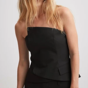 Korsett  - Den här bandeautoppen har en asymmetrisk, axelbandslös korsettdesign och en överlappande stil. Den har en inre silikonkant runt bröstet, ett resårband på baksidan med ett fiskekrokspänne och en falsk framficka med lock. Liten i storleken. Oanvänd 