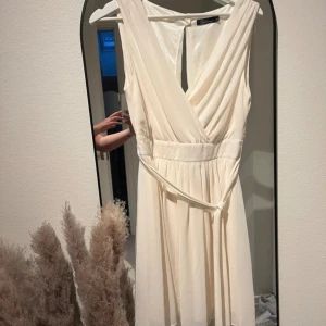 Champagnefärgad klänning med omlott - Säljer en elegant champagnefärgad klänning med omlott-design. Klänningen är ärmlös och har en vacker v-ringning. Perfekt för fest eller sommarens alla tillställningar. Den är i nyskick och har en normal passform. Materialet känns mjukt och skönt mot huden.
