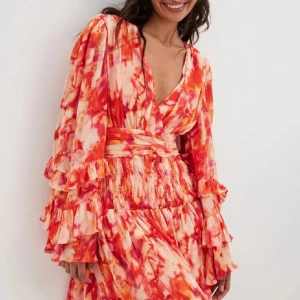 Färgglad kortklänning med volanger - Säljer en superfin och färgglad kortklänning med volanger. Klänningen har ett mönster i rött, orange och vitt som verkligen sticker ut. Den har långa ärmar med volangdetaljer och en v-ringad hals. Perfekt för sommarens alla fester! 💃