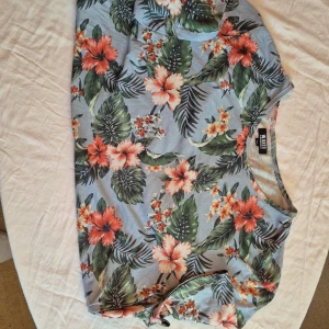 Blommig t-shirt från William Baxter - Säljer en snygg blommig t-shirt från William Baxter i storlek XL. T-shirten har ett färgglatt mönster med gröna blad och orangea blommor på en ljusgrå bakgrund. Perfekt för sommaren och att ha på fest! 🌺