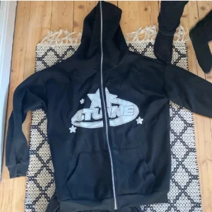 Svart hoodie med dragkedja och matchande byxor - Säljer en svart hoodie med dragkedja och matchande byxor. Hoodien har ett stort tryck på framsidan med texten 'ATUNE' och en stjärna. Byxorna har ett matchande tryck med texten 'NAPS' och en stjärna. Perfekt för en avslappnad dag eller träning. Båda plaggen är i bra skick och väldigt bekväma.