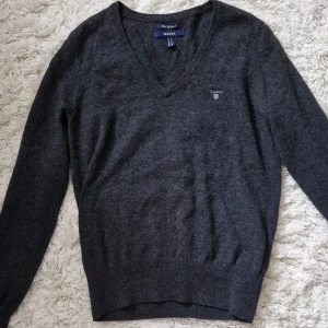 Mörkgrå tröja från GANT - Säljer en snygg och stilren mörkgrå tröja från GANT. Tröjan är i storlek M och har en v-ringad hals. Den är perfekt för både vardag och lite finare tillfällen. Tröjan har långa ärmar och en liten broderad GANT-logga på bröstet. Passar perfekt till höst och vinter!