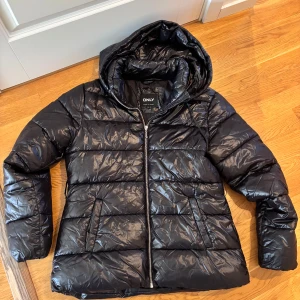 Svart pufferjacka från ONLY - Säljer en snygg svart pufferjacka från ONLY. Den är perfekt för kalla dagar med sin vadderade design och har en praktisk huva. Jackan har en dragkedja framtill och är i mycket bra skick. Perfekt för både höst och vinter!