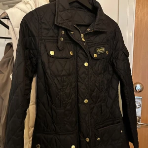Svart quiltad jacka från Barbour - Säljer en snygg svart quiltad jacka från Barbour. Den har guldiga knappar och en dragkedja framtill. Jackan är perfekt för höst och vår, med en stilren design och praktiska fickor. Den är långärmad och har en normal passform.