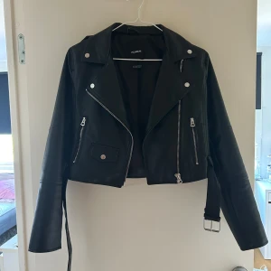 Svart skinnjacka från Pull&Bear - Säljer en snygg svart skinnjacka från Pull&Bear. Jackan har en cool bikerstil med dragkedjor och knappar som detaljer. Den är perfekt för höst och vår och ger en edgy look till vilken outfit som helst. Jackan är i storlek M och har långa ärmar.