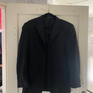 Svart kavaj med ullblandning - Säljer en klassisk svart kavaj från Pendleton i storlek 46. Den är tillverkad i en ullblandning med 60% ull och 2% elastan för extra stretch och komfort. Perfekt för både formella tillfällen och vardagsbruk. Kavajen har en stilren design med knappar framtill.