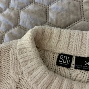 Off-white stickad tröja från BDG Urban Outfitters - Säljer en superfin off-white stickad tröja från BDG Urban Outfitters. Den har ett snyggt kabelstickat mönster och är perfekt för både höst och vinter. Tröjan är i storlek S och har en normal passform. Den är i nyskick. Nypris 899kr