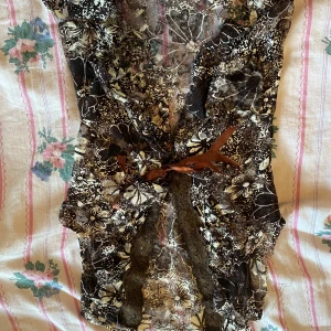 Spetts top - Säljer en superfin blommig lace topp med spetsdetaljer och en söt rosett framtill. Toppen har ett unikt mönster i svart, beige och bruna toner. Perfekt för en festlig kväll eller en dag när du vill känna dig extra speciell!