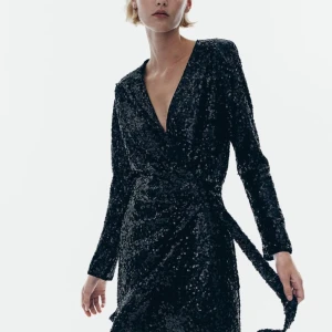 Svart glittrig klänning med knytband - Snygg svart klänning med glittriga paljetter och v-ringad design. Klänningen har långa ärmar och ett knytband i midjan för en smickrande passform. Perfekt för festliga tillfällen.