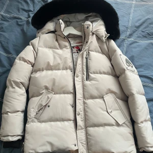 Moose knuckles jacka - Snygg beige dunjacka från Moose Knuckles med svart pälsdetalj på huvan. Jackan har dragkedja och knappar framtill samt flera praktiska fickor. Perfekt för kalla vinterdagar. Liten defekt knappen längst upp har tråkigt nog lossnat. Jackan är strl L men sitter som M. Priset är inte hugget i sten. MVH/ Liam
