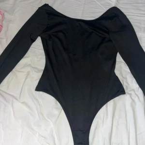 Svart långärmad bodysuit - Snygg svart långärmad bodysuit med öppen rygg i 100% polyester. Perfekt för en stilren look med en bekväm passform. Bodysuiten har en praktisk knäppning i grenen. Helt ny oanvänd 