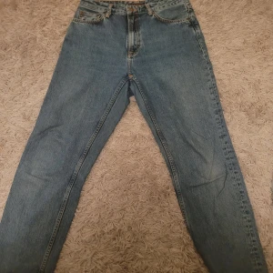 Blå jeans från Nudie - Snygga blå jeans från Nudie med klassisk femficksdesign och knappgylf. Jeansen har en normal passform och är perfekta för en avslappnad stil. Detaljerad söm på bakfickan ger en extra touch.