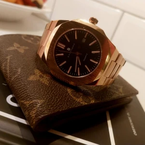 Guldfärgad klocka med svart urtavla - Snygg guldfärgad klocka med svart urtavla och datumvisning. Armbandet är i metall med en klassisk länkdesign. Perfekt accessoar för en stilren look.