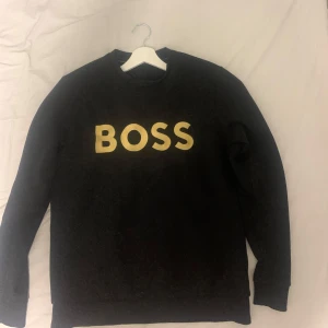  Boss Sweatshirt  - Snygg svart tröja från Boss med stort gult tryck på framsidan. Perfekt för en stilren och avslappnad look. Tröjan har långa ärmar och en klassisk passform.