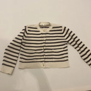 Randig kofta från Gina Tricot - Säljer en snygg randig kofta från Gina Tricot i storlek 146/152. Koftan är i beige och svart med guldknappar framtill. Perfekt för en stilren look!