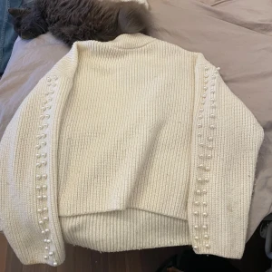 Vit stickad tröja med pärldetaljer - Säljer en stilren vit stickad tröja med pärldetaljer längs ärmarna. Tröjan har en klassisk design med ribbade kanter och långärmad passform. Perfekt för att ge en elegant touch till din outfit.