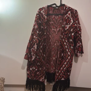 Mönstrad kimono med fransar - Snygg och unik kimono i rött, svart och vitt med ett iögonfallande mönster. Den har trekvartsärmar och fransar längs nederkanten som ger en bohemisk touch. Perfekt för att piffa upp vilken outfit som helst.