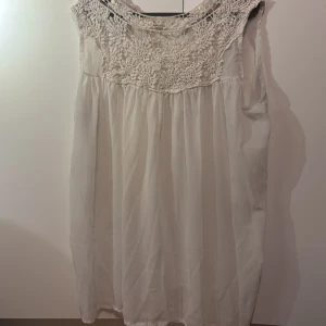 Vit ärmlös blus med spetsdetaljer - Elegant vit blus med vacker spetsdetalj vid halsringningen. Blusen är ärmlös och har en luftig passform, perfekt för varma dagar. Den romantiska spetsen ger en feminin touch.