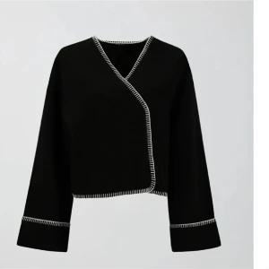 Svart omlott kofta med vita detaljer - Snygg svart omlott kofta med vita sömmar längs kanterna. Koftan har en croppad passform och långa ärmar, perfekt för en stilren look.