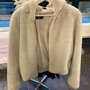 Beige teddyjacka från ONLY - Mysig beige teddyjacka från ONLY med dragkedja framtill. Jackan har en fluffig textur och långa ärmar, perfekt för kyligare dagar. Den är i en normal passform och ger en trendig look.