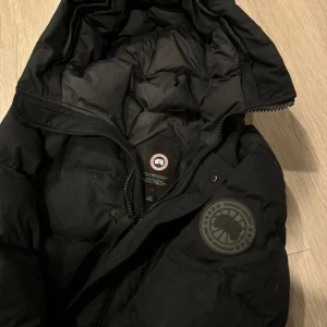 Svart dunjacka med huva - Säljer en svart dunjacka från Canada Goose med huva och dragkedja. Jackan har en rund logotyp på ärmen och är perfekt för kalla dagar. Den är både stilren och funktionell.