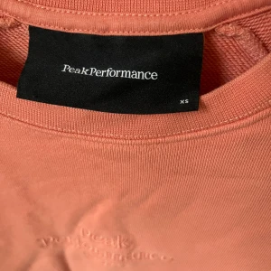 Melon färga tröja från Peak Performance - Säljer en snygg  tröja från Peak Performance i storlek XS. Tröjan har en broderad logga på bröstet och långa ärmar. Perfekt för en avslappnad stil. Tröjan aldrig använd behöv bara att strykas. 