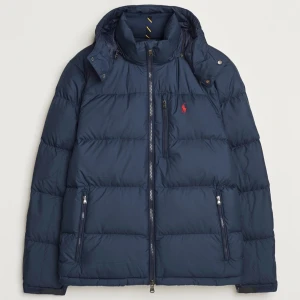 Blå dunjacka från Polo Ralph Lauren - Snygg blå dunjacka från Polo Ralph Lauren med dragkedja och avtagbar huva. Jackan har en broderad röd logotyp på bröstet och praktiska fickor med dragkedja. Perfekt för kyliga dagar.