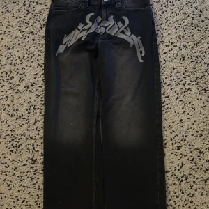 Vicinity Jeans - Coola svarta jeans med unikt tryck på framsidan. De har en klassisk femficksdesign och en vit logotyp på baksidan av midjan. Perfekta för en avslappnad stil med en twist.