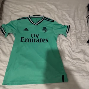 Real Madrid tröja - Snygg grön fotbollströja från Adidas med korta ärmar och svart text. Tröjan har en klassisk krage med svarta detaljer och klubbmärke på bröstet. Perfekt för fotbollsfans! Pris kan diskuteras
