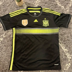 Fotbollströja/Jersey Spanien FIFA 2010 world cup - Snygg svart fotbollströja från Adidas med gula detaljer och korta ärmar. Tröjan har ett emblem på bröstet och tre gula ränder på axlarna. Perfekt för fotbollsfans! Riktigt god kvalité för hur mycket tid det har gått, ganska svårt att hitta liknande. 