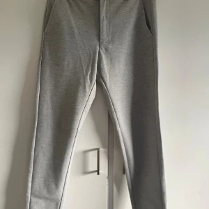 Grå byxor från Lindbergh - Snygga grå byxor från Lindbergh i slim fit och croppad längd. Perfekta för en stilren look med knappdetalj i midjan. Passar bra till både vardag och mer uppklädda tillfällen.