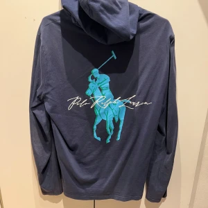 Polo Ralph lauren hoodie - Säljer en snygg mörkblå hoodie från Polo Ralph Lauren med en stilren logga i ljusblått på ryggen och ett stort tryck på bröstet. Perfekt för en avslappnad stil. Lampan gör så den ser lite smutsig ut men det är bara lampan!