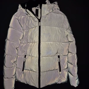 Reflekterande pufferjacka med huva - Snygg reflekterande pufferjacka med huva och dragkedja framtill. Jackan har två fickor med dragkedjor och är perfekt för kalla dagar. Den har en modern look och är både praktisk och stilren.