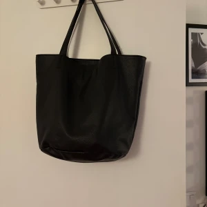 Svart axelväska i skinn - Snygg och rymlig svart axelväska i skinn med enkel design. Perfekt för att bära med sig allt man behöver under dagen. Väskan har två handtag och en stilren look.