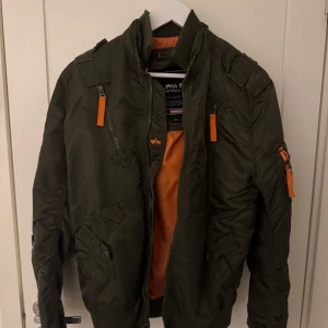 Grön bomberjacka från Alpha Industries - Säljer en grön bomberjacka från Alpha Industries med orange foder och flera dragkedjor. Jackan har broderade detaljer på ärmarna och en klassisk bomberstil. Perfekt för höst och vår. Nypris: 2000-3000kr 