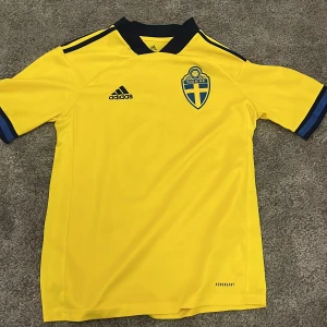 Gul fotbollströja från Adidas Sverige - Säljer en gul fotbollströja från Adidas med Sveriges landslagsemblem på bröstet. Tröjan har korta ärmar och mörkblå detaljer vid krage och i slutet av ärmarna. Är för storlek XL för barn passar ca 160 cm. Tror den är från 2020. Nypris var ca 1000 