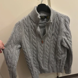 Grå stickad tröja från Polo Ralph Lauren - Säljer en grå stickad tröja från Polo Ralph Lauren med kabelstickat mönster och dragkedja vid halsen. Tröjan är helt ny haft på mig den en gång. Säljer den för att den kändes för tight på armarna, så passar inte för folk som har stora biceps.