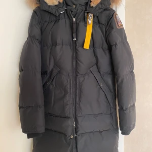 Svart dunjacka med päls från Parajumpers - Snygg svart dunjacka från Parajumpers med pälsfodrad huva och ikonisk gul rem. Jackan har dragkedja och knappar framtill samt flera praktiska fickor. Perfekt för kalla vinterdagar.