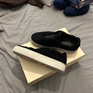 Svarta sneakers från Axel Arigato - Snygga svarta sneakers från Axel Arigato med en stilren design. Skorna har en vit sula och är tillverkade i mocka, vilket ger dem en lyxig känsla. Perfekta för en avslappnad men stilfull look. Arigatopåse och skolåda följer med