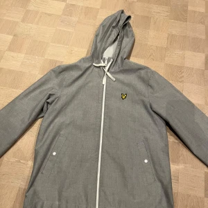 Grå jacka med huva från Lyle & Scott - Snygg grå jacka med huva från Lyle & Scott. Jackan har en dragkedja framtill och två fickor med knappar. Perfekt för vår och höst. Mycket bra skick har blivit använd 1-2 gånger.