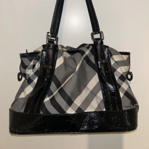 Burberry Handväska - Äkta Burberry Handväska i svart/grått/vitt. Vintage. Modellen heter ”Beat Check Lowry” - canvas tote bag. Väska har lite slitningar på sidorna och fläckig på undersidan. Jag kan skicka bättre bild på detta om det är svårt att se på sista bilden! Annars i fint skick! Hör av er vid frågor/foer bilder!💓 (Har tyvärr inga orginalförfackningar, men ett kort tillkommer)