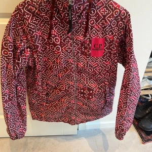 Mönstrad jacka från C.P. Company - Säljer en unik mönstrad jacka från C.P. Company i rött och vitt. Jackan har en cool design med huva och inbyggda glasögon, perfekt för att sticka ut. Den har dragkedja framtill och oanvänd. Perfekt för höst och vår!