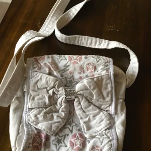 Axelväska med rosett - Charmig beige axelväska med quiltad design och en söt rosett framtill. Väskan har ett justerbart axelband och en dekorativ broderad detalj i olika färger på locket. Perfekt för att ge en romantisk touch till din outfit.