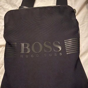 Svart axelväska från Hugo Boss - Snygg svart axelväska från Hugo Boss med justerbar rem och dragkedja. Perfekt för att bära dina dagliga nödvändigheter med stil. Väskan har en diskret logotyp på framsidan och är tillverkad i ett slitstarkt material.