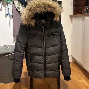 Svart dunjacka med päls från Lindberg - Snygg svart dunjacka från Lindberg med avtagbar huva och pälsdetaljer. Jackan har dragkedja och flera praktiska fickor med dragkedjor. Perfekt för kalla vinterdagar.