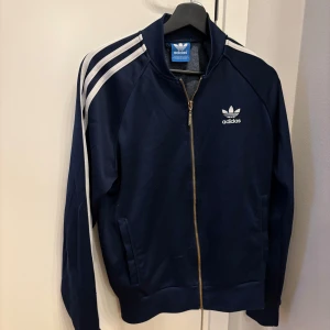 Mörkblå träningsjacka från Adidas - Snygg mörkblå träningsjacka från Adidas med klassiska vita ränder längs ärmarna och dragkedja framtill. Jackan har en broderad logga på bröstet och långa ärmar. Perfekt för en sportig look!
